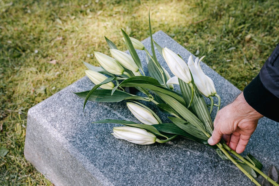 A bouquet of white lilies with long green leaves, arranged on a grey stone gravestone, being gently placed or adjusted by a person wearing a dark sleeve. The lilies are fresh, with closed buds and ope