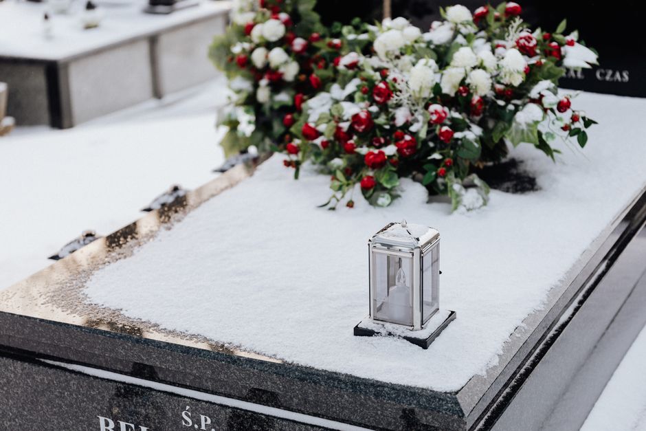 A floral tribute featuring a lush spray of fresh white carnations, red berries, and green foliage arranged on a snow-covered stone surface at a burial site. The bouquet is partially visible, resting o