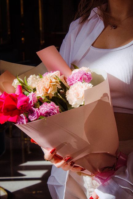 A person holding a beautifully arranged bouquet of fresh flowers wrapped in beige paper. The bouquet features pink, white, and peach carnations, along with vibrant pink spray roses and greenery. The f