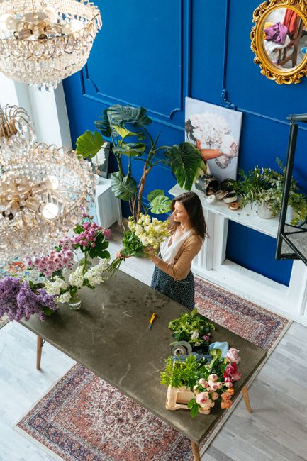 A woman arranging a floral bouquet inside a brightly lit floral studio with deep blue walls. She is holding a large bouquet of fresh white roses, pink carnations, and greenery, preparing a floral gift
