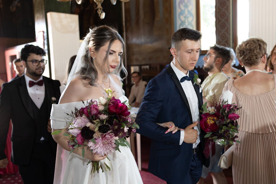 A bride and groom stand indoors during a wedding celebration, each holding a large floral bouquet featuring deep pink peonies, blush pink dahlias, dark purple anemones, and sprigs of greenery, wrapped