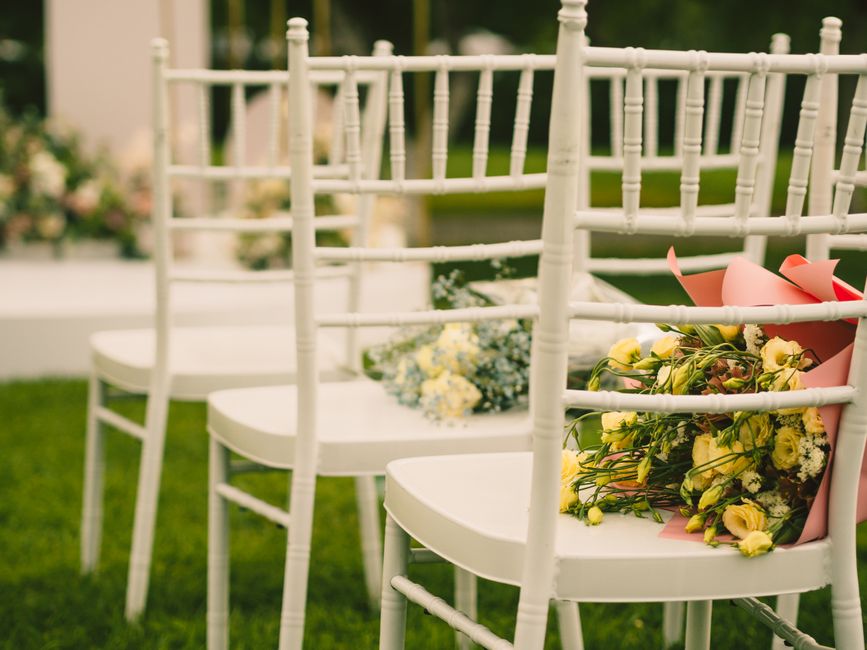 A row of white wooden chairs arranged outdoors on a lush green lawn, each adorned with floral arrangements. The bouquet on the nearest chair features yellow and cream roses, interspersed with white ba