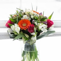 This lavish bouquet is a testament to floral artistry, combining brilliant colour with intricate structure-all grounded in the character of Streatham. At the heart, peach and orange gerbera daisies create a glowing halo, each petal echoing the soft pastel dawns over Streatham Common and the bustling energy of the High Road. Deep fuchsia roses add a layer of drama and romance, their petals tightly wound in perfect spirals that suggest both passion and contemplation. Ivory-white alstroemeria, with their fine, multi-petaled heads and green-flecked throats, weave in a brightness that lifts the entire display, while tiny white and blush filler flowers lend delicacy and movement. Around the base, broad aspidistra leaves are carefully curved to form a protective green collar-reminiscent of the leafy calm in The Rookery's historic gardens-offering structure and a sense of completion. Tapered greenery and feathery sprigs fill any spaces, ensuring every angle is as lush and generous as the last. The arrangement rests in a tall, clear glass vase where the vibrant green stems seem to reach downward for water, accentuating the design's fresh-cut vitality. Soft, diffuse light brings out the bouquet's depth and subtle gleam, while a fleeting fragrance-part garden, part spring morning-rises from the petals. This arrangement feels tuned to the rhythms of Streatham life: perfect for joyful gatherings, marking milestones, or brightening a friend's day across SW16. Each element is chosen and placed with the innate understanding of a neighbourhood florist deeply rooted in the local community.